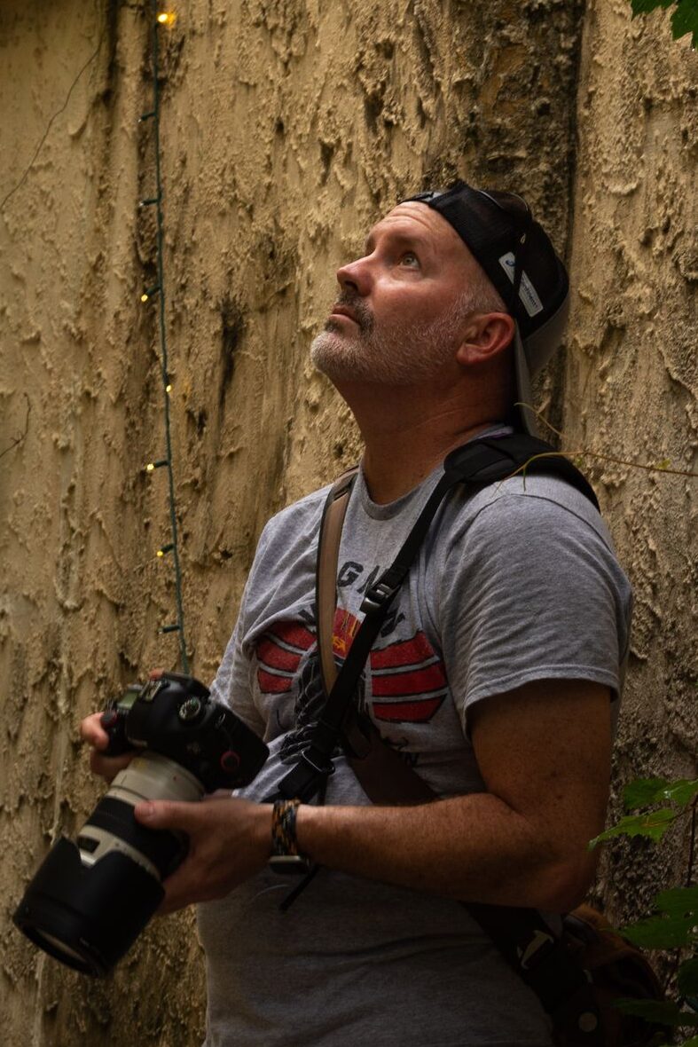 A portrait of David wearing a baseball cap backwards, leaning against a textured stucco wall, holding a digital SLR camera, looking up towards the sky.